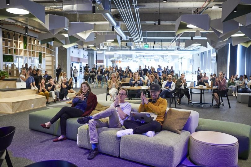 A group of Atlassian employees in a modern office with couches, applauding a teammate. 