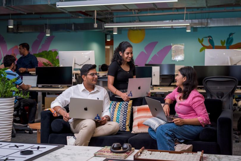 Atlassian employees chat with one another on a couch, laptops in hand, in front of a colorful mural